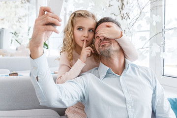 daughter covering eyes of father and showing silence sign while they taking selfie
