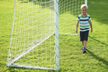 Cute little blond boy playing at being a goalkeeper on a sportsfield standing in the goalposts in sunny summer day
