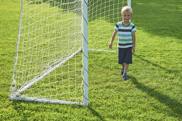 Cute little blond boy playing at being a goalkeeper on a sportsfield standing in the goalposts in sunny summer day
