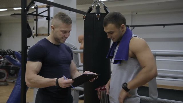 Young sportsman is discussing training results his instructor in the gym. Athlete with blue towel is drinking water from the bottle and his male trainer is looking to papers on the notepad in his
