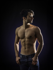 Fototapeta na wymiar Handsome, fit muscular young man shirtless, wearing only jeans standing on black background, looking away to a side