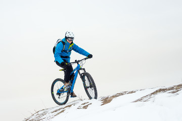 Fototapeta premium Cyclist in Blue Riding Mountain Bike on Rocky Winter Hill Covered with Snow. Extreme Sport and Enduro Biking Concept.