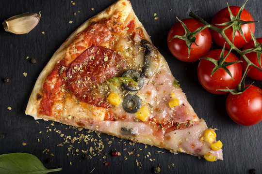 Slice of Italian Capriciosa pizza on dark slate with dried oregano