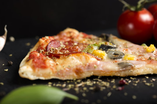 Slice of Italian Capriciosa pizza on dark slate with dried oregano