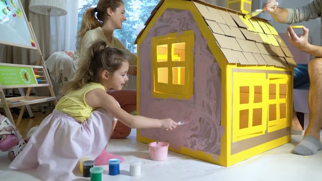 Young family with little girl building and painting toy cardboard house together.