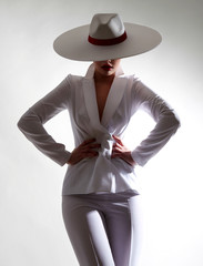 Beautiful young girl with a beautiful figure in a white trouser suit and a wide-brimmed white hat posing against a white background in the studio.