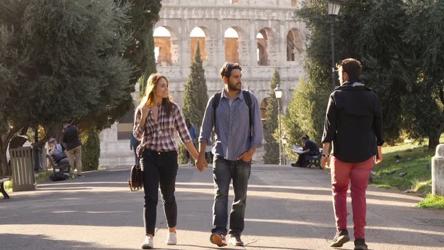 Young couple walks in park road holding hands when attractive man glances at girlfriend. She gets distracted and boyfriend gets jealous. Disoloyal girl and envy. Colosseum in the background in rome at