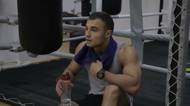 Portrait of young male pugilist, who is resting after the sparring in boxing gym. Sportsman with tattoo in grey sleveless t-shirt is sittning near the ring and punching bag, drinking water and