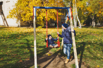 Young woman walk in green yellow fall autumn park with little cute child baby boy on nature. Mother, kid son fun rest, ride on swing. Parenthood, family day 15 of may, love, parents, children concept.