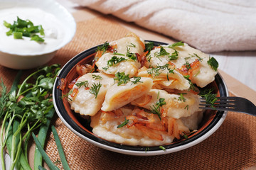 A plate with boiled dough vareniki with cabbage filling and fried onions. Near her bowl with sour cream, fork and greens.