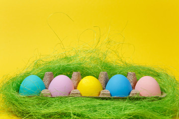 Row of five colorful pastel monophonic painted Easter eggs in cardboard tray in green grass on yellow background for card. Happy Easter concept. Copy space for advertisement. With place for text.