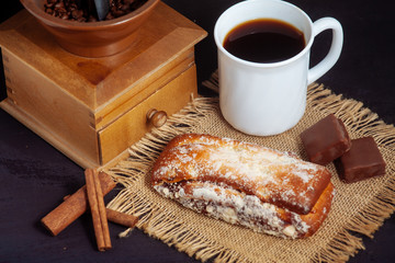A cup of coffee with a cake, with chocolate sweets and an old hand grinder on a black textured table.