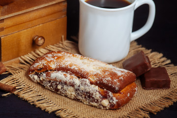 A cup of coffee with a cake, with chocolate sweets and an old hand grinder on a black textured table.