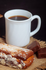 A cup of coffee with a cake, with chocolate sweets and an old hand grinder on a black textured table.