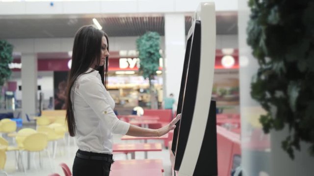 Caucasian female using automated teller machine with big digital screen while standing in shopping mall, woman verifies account balance on banking application via modern device icon