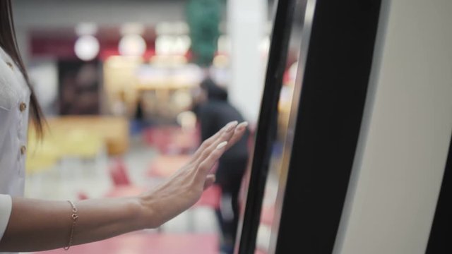 Caucasian female using automated teller machine with big digital screen while standing in shopping mall, woman verifies account balance on banking application via modern device icon