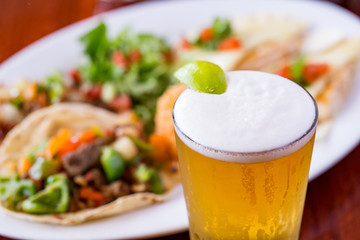 Cold beer with lime wedge and Mexican food dish in beef tacos in background.