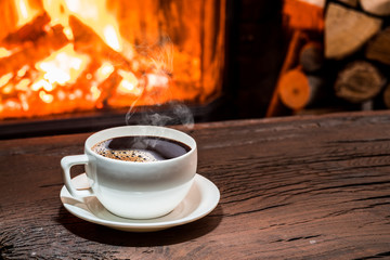 Cup of hot coffee on the wooden table and fireplace at the background.