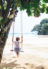 Fashionable dressed Woman sits on tree swing on the wide sandy ocean beach