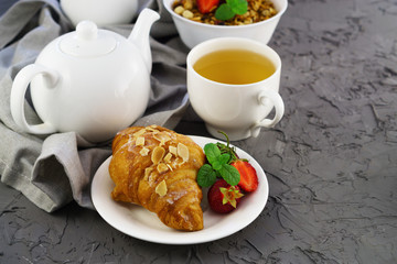 Delicious continental or French breakfast concept. Fresh flaky croissants, cereal, mint, strawberry and green tea in white cup and teapot on gray cement table. Close up, selective focus, copy space.