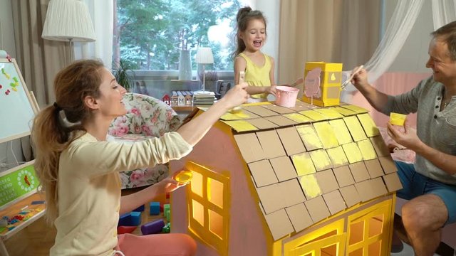 Young family with little girl building and painting toy cardboard house together. Slow motion-
