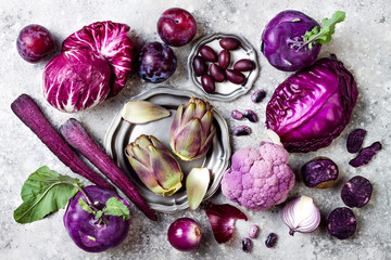 Raw purple vegetables over gray concrete background. Cabbage, radicchio salad, olives, kohlrabi, carrot, cauliflower, onions, artichoke, beans, potato, plums. Top view, flat lay.