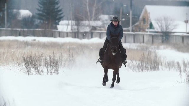 Woman in cawboy hat galloping on big dark horse in snow field - slow-motion