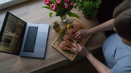 The pregnant girl is watching the show and preparing salmon with rosemary and lemon for lunch. on the table are fresh tulip flowers