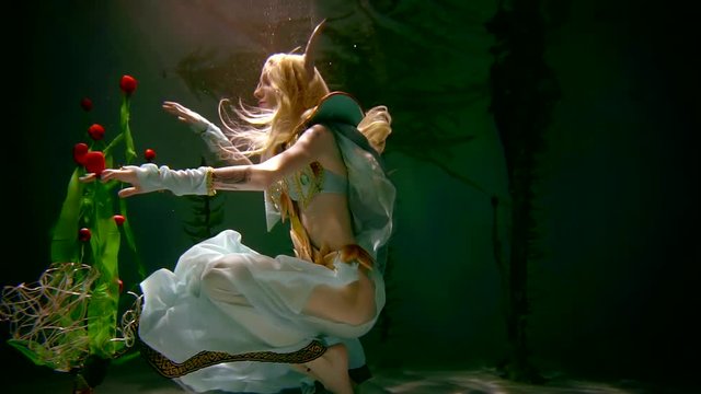 Engaging woman in elf costume is posing underwater with many flowers. She is reaching tulips with her hand. Reflection on the surface.