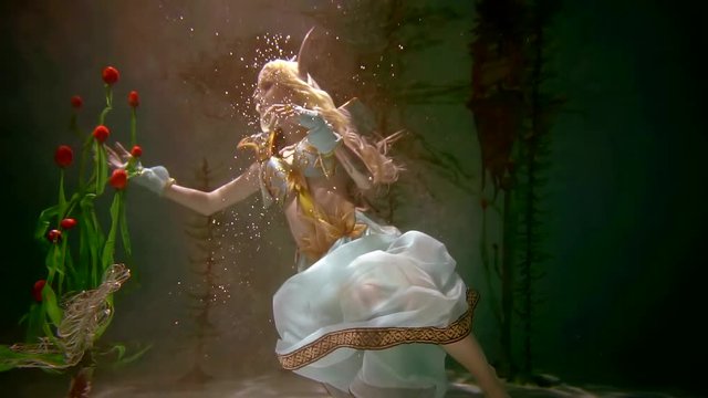 Engaging woman in elf costume is posing underwater with many flowers. She is reaching tulips with her hand. Reflection on the surface.