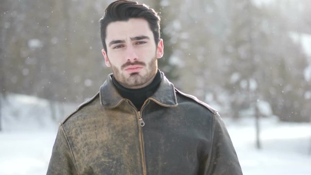 Handsome young man posing among snowy woods in the mountain pretending to shoot a bullet to his head with gun made with fingers, wearing leather winter jacket