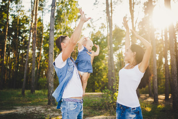 Obraz premium Theme family vacation in the forest. A small child has daughter with daddy on shoulders, mother stands next to her raised arms and smile. Dad and mom playing with small child. Wearing jeans clothes