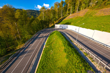 A winding modern asphalt road to the top of a resort in the bright yellow rays of the evening sun, dense forest and the Caucasian mountain range. Summer spring forest mountain landscape, Sochi Russia.