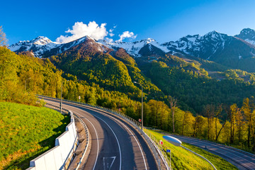 Fototapeta na wymiar A winding modern asphalt road to the top of a resort in the bright yellow rays of the evening sun, dense forest and the Caucasian mountain range. Summer spring forest mountain landscape, Sochi Russia.