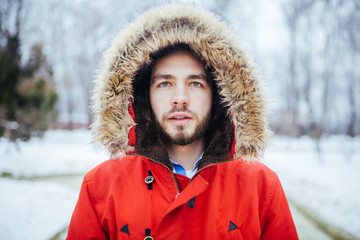 Portrait, close-up of a young stylish man with a beard dressed in a red winter jacket with a hood and fur on his head stands against the background of a snow-covered city. Winter and frost theme