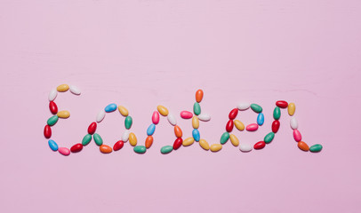 Colorful egg-shaped, sugar coated Easter candy in rainbow colors filled with meringue, chocolate or bubble gum laid out on light pink wooden background saying the word easter