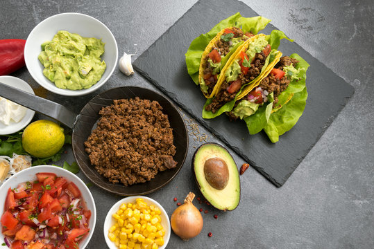 stuffed mexican tacos and ingredients like fried ground beef, tomato salsa, guacamole, corn and spices on a dark slate plate, top view from above