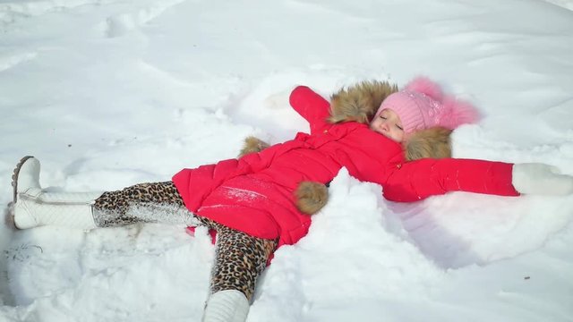 Portrait of a beautiful young girl laying down on a frozen snow lake moving her arms and legs up and down creating a snow angel figure, playing games during a sunny winter vacation.