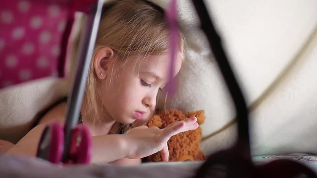 Cute little girl sitting on bed together with teddy bear playing games on tablet.