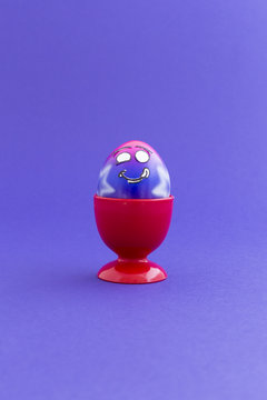 Pink and purple painted Easter egg with funny cartoon style face in a red plastic egg cup and purple background