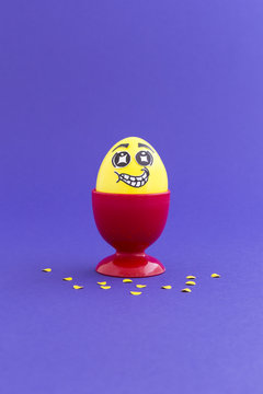 Yellow painted Easter egg with funny cartoon style face in a red plastic egg cup and yellow confetti on purple background