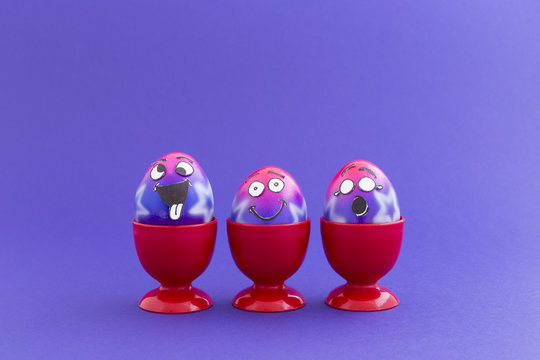 Group of colorful painted Easter eggs with funny cartoon style faces in red plastic egg cups on purple background