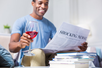 Alcoholic drink. Selective focus of a glass with red wine being in hands of a cheerful nice positive man while reading a newspaper