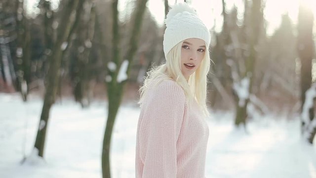 Young pretty blonde girl in white hat and pink sweater walking in the winter forest. Girl goes to the camera, fixes her hair and then dances.
