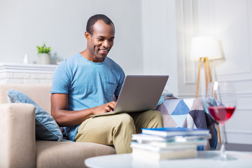 Modern entertainment. Happy joyful nice man smiling and using laptop while sitting on the sofa