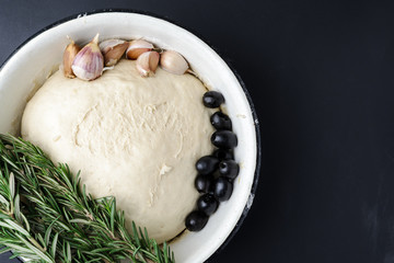 Raw dough in a white bowl, sprigs of rosemary, olives and garlic on a dark background.