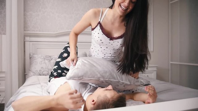 Happy cheerful loving couple having a pillow fight in bed, young attractive guy and girl sitting in pajamas, slow-motion