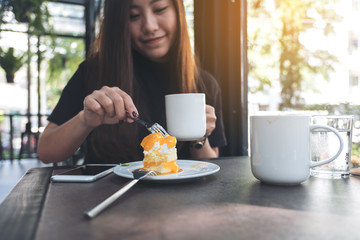 Closeup image of a beautiful Asian woman eating an orange cake while drinking coffee in modern cafe