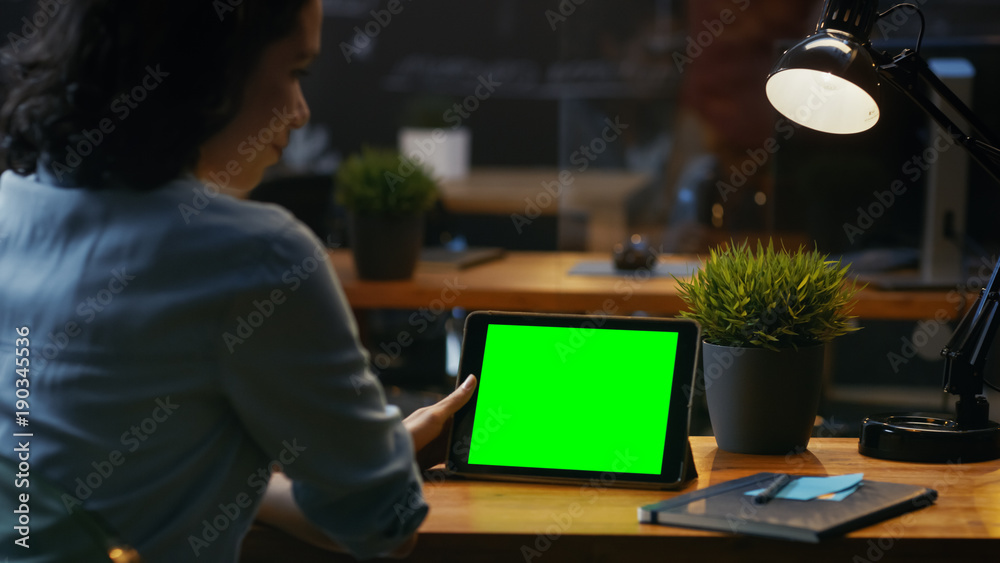 Poster young woman sits at her wooden desk uses tablet computer with mock-up green screen, she uses various