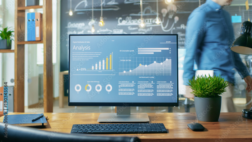 Poster Personal Computer with Mobile Application Design Showing on the Monitor Stands on the Office Desk, In the Background Man Working in the Daytime Office Environment.